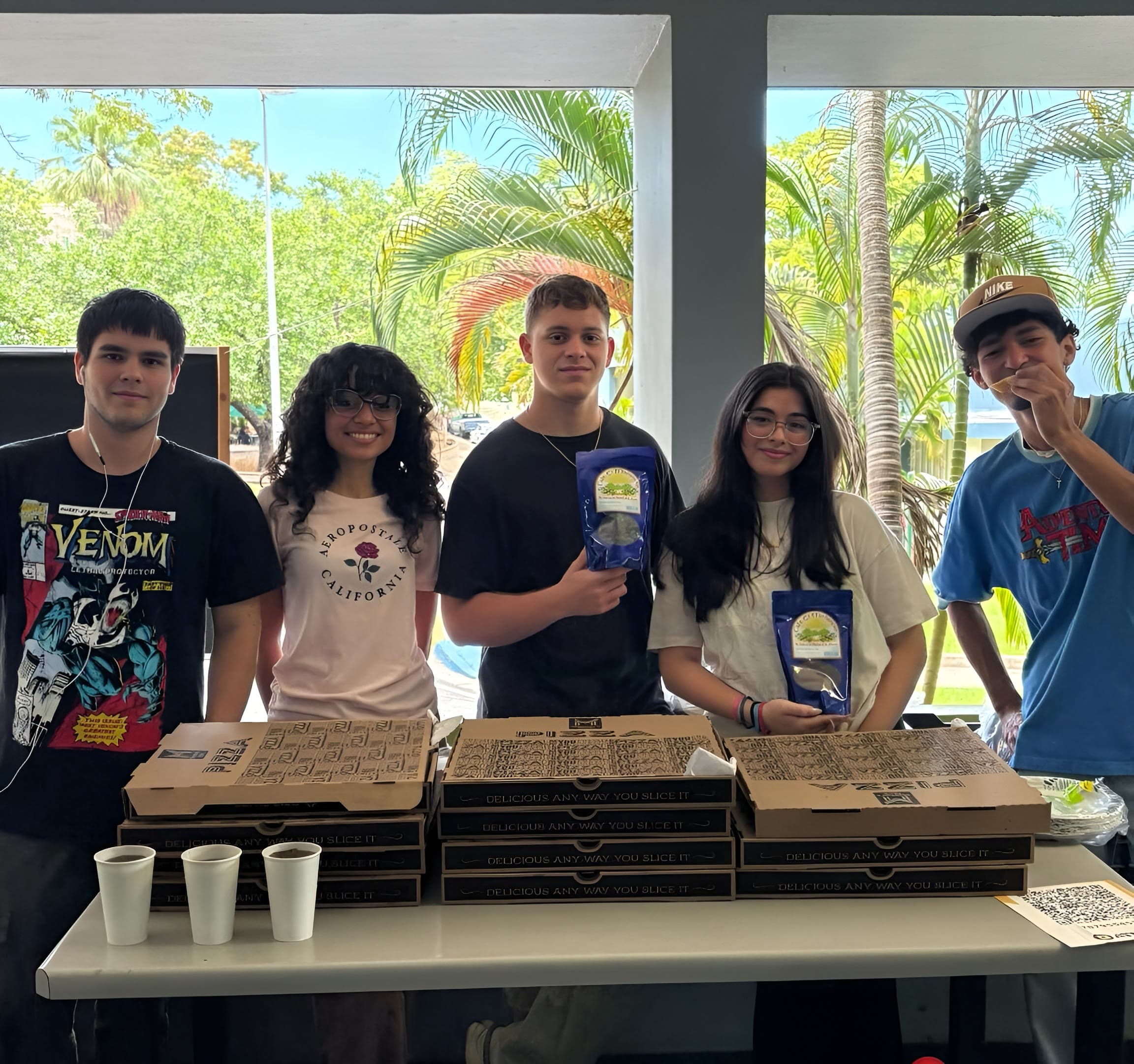 Five DRACO members standing behind a table stacked with pizza boxes and bags of coffee during a campus fundraiser