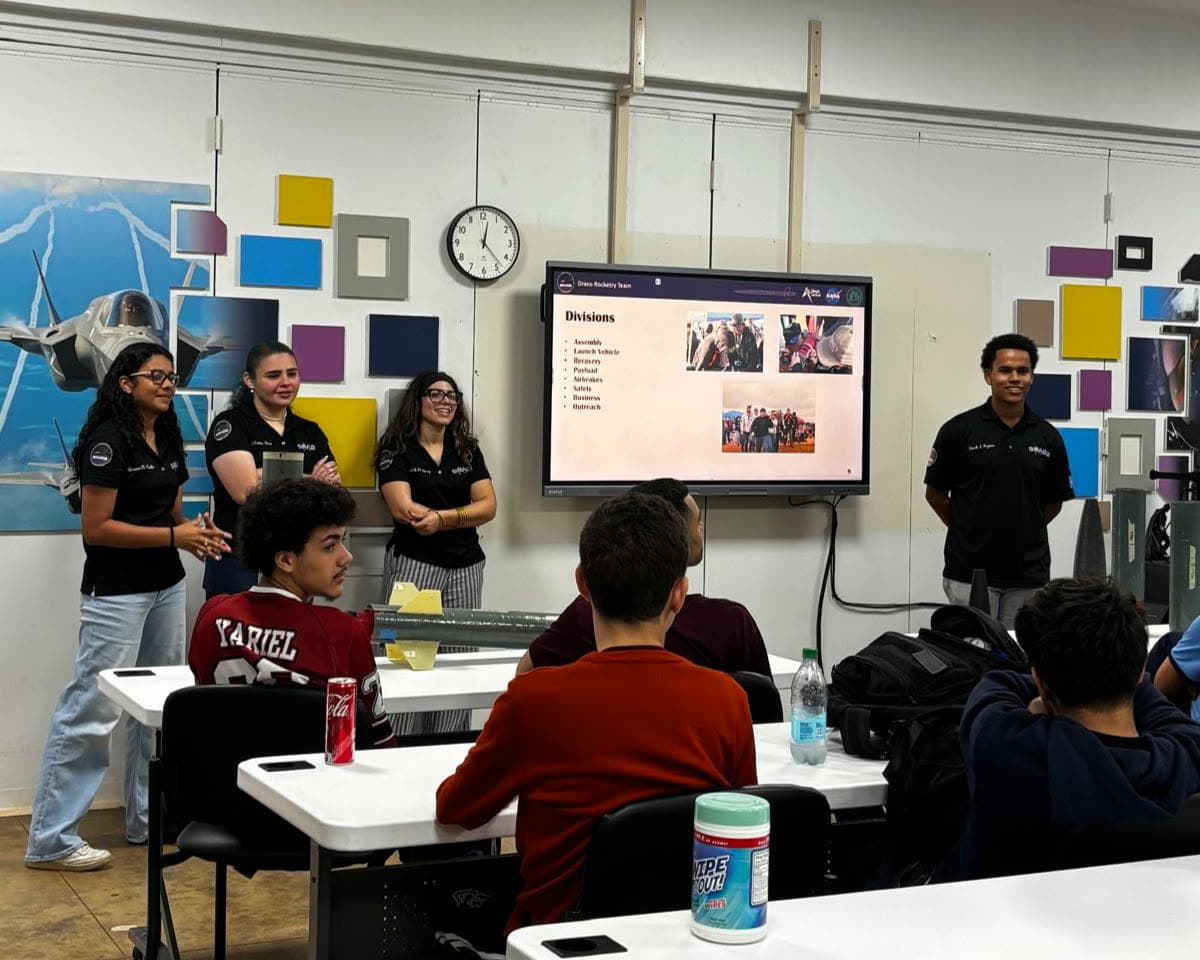 Four DRACO members presenting the team's divisions to high-school students, with rocket hardware on the desk in front of them