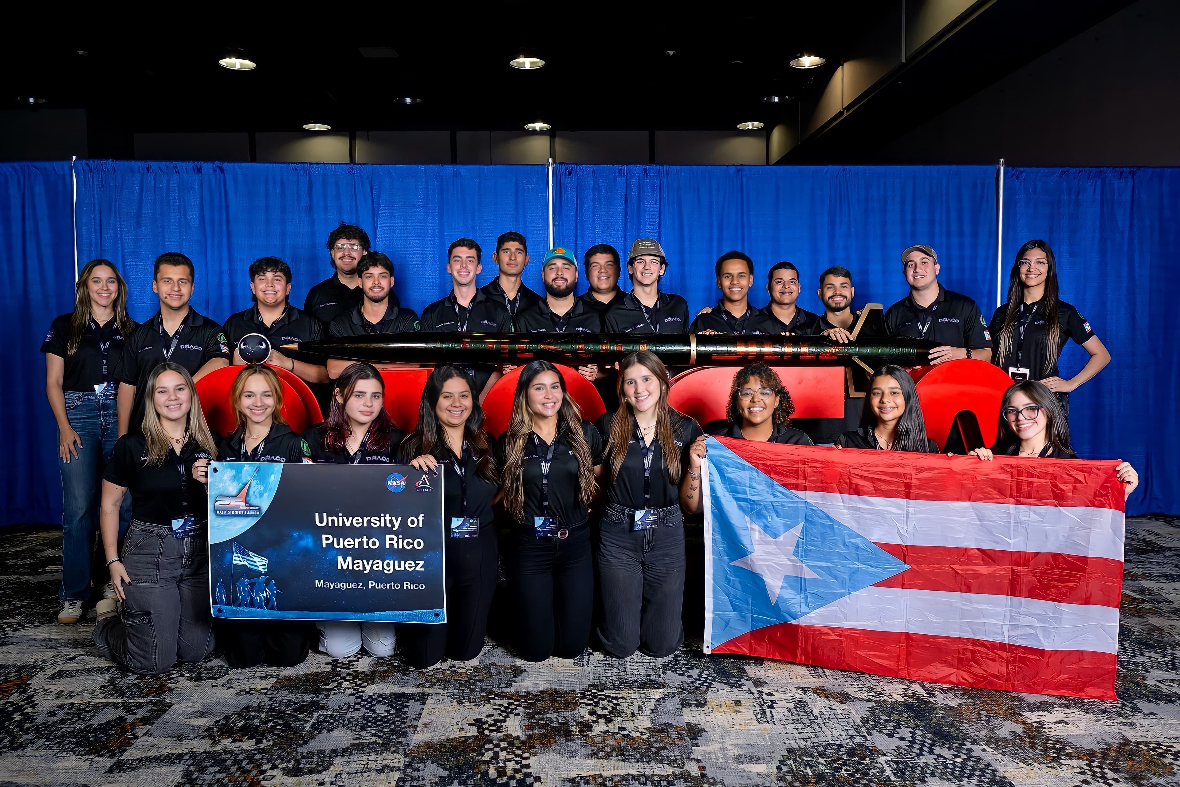 The DRACO team posed with the Forest Wyvern rocket, a University of Puerto Rico at Mayagüez sign, and a Puerto Rico flag at the 2025 NASA Student Launch competition venue