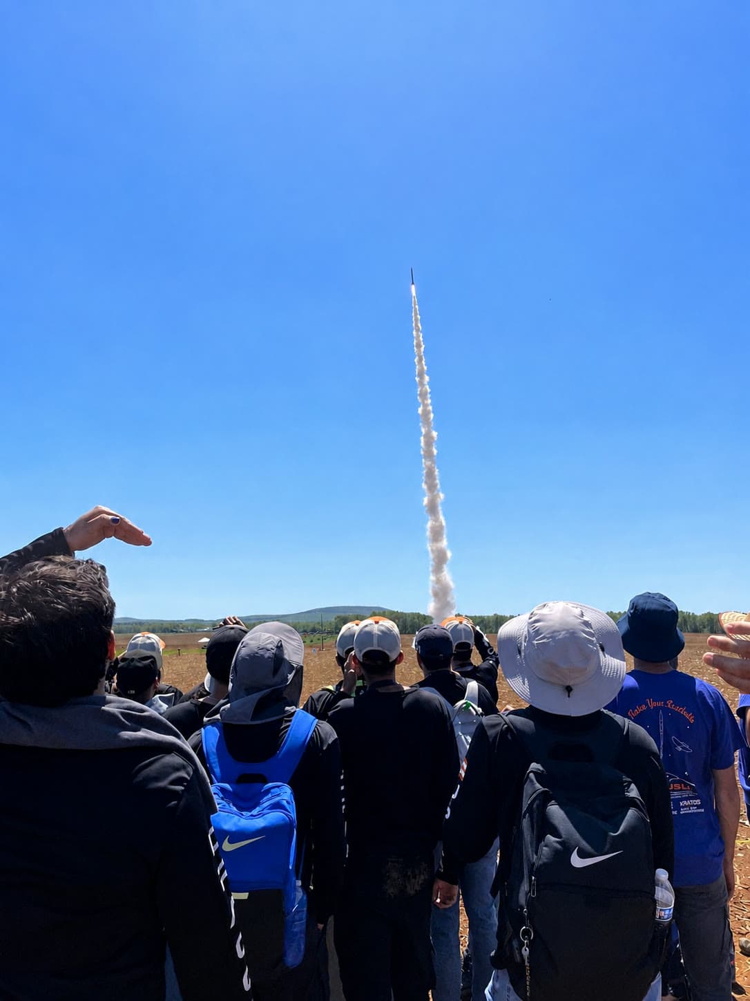 A DRACO rocket climbing into a clear blue sky as the team watches from below