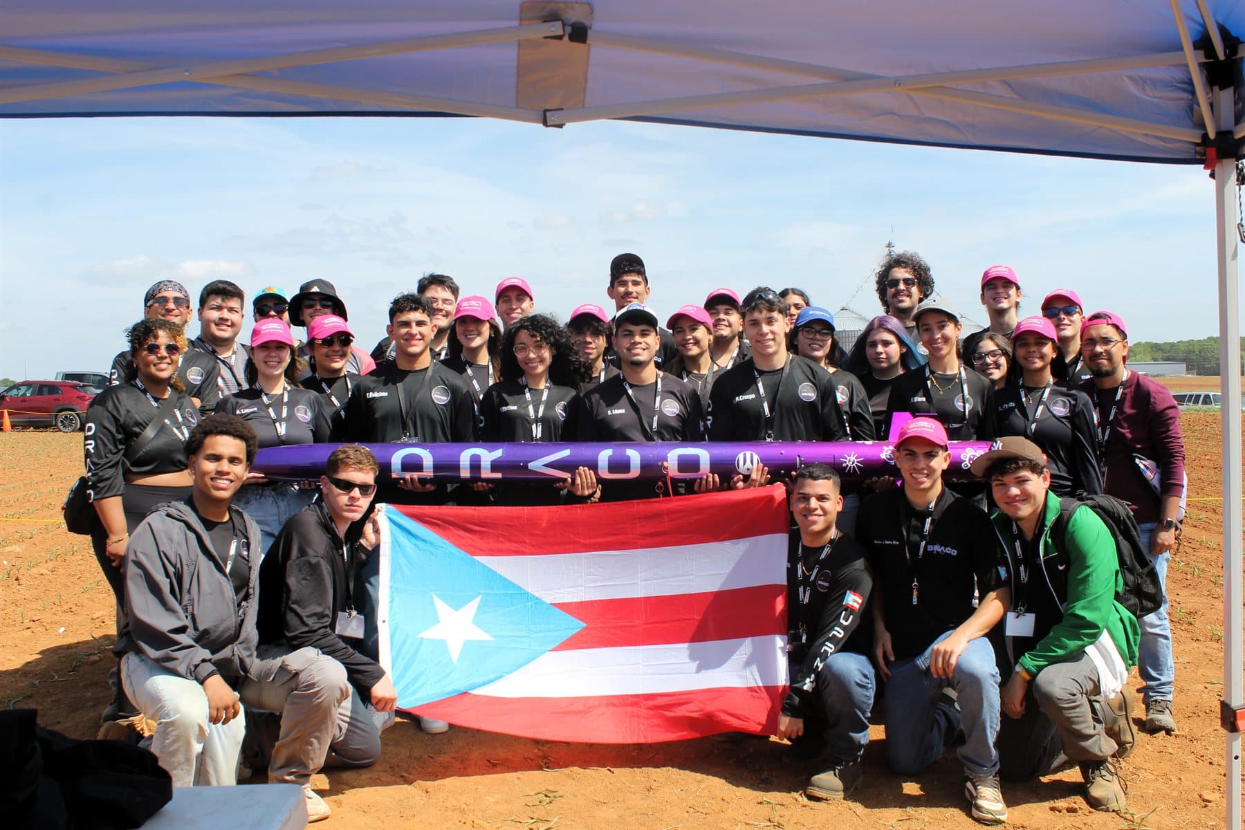 The 2026 DRACO team gathered under a canopy at the launch field, holding the purple Koeia airframe and a Puerto Rico flag