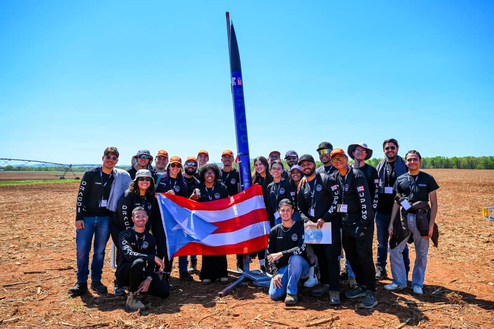 The 2024 DRACO team standing in a field beside the blue Starfire rocket on its launch rail, holding a Puerto Rico flag under a clear sky